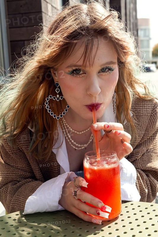 A woman with stylish accessories, including large hoop earrings and layered necklaces, sips a refreshing red drink outdoors on a sunny day, exuding a casual chic vibe