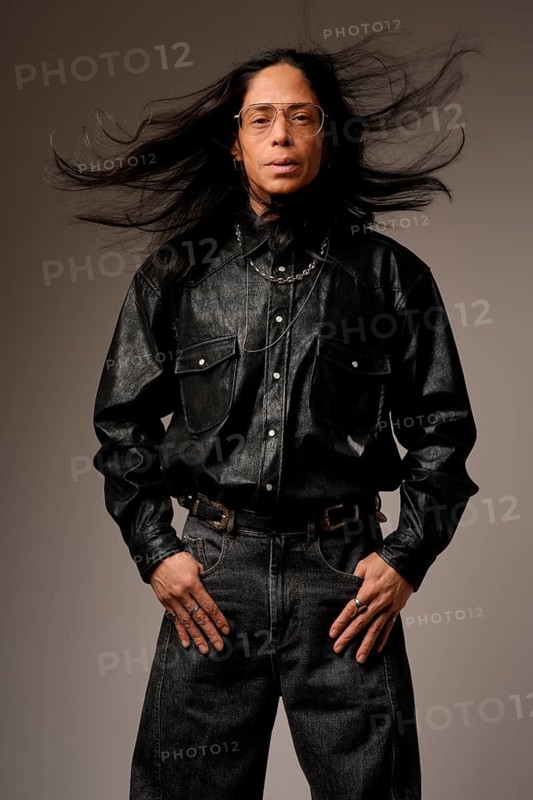 Portrait of a Latin man wearing glasses and a black leather shirt. His long hair is dramatically blown by the wind, adding dynamic movement to the image