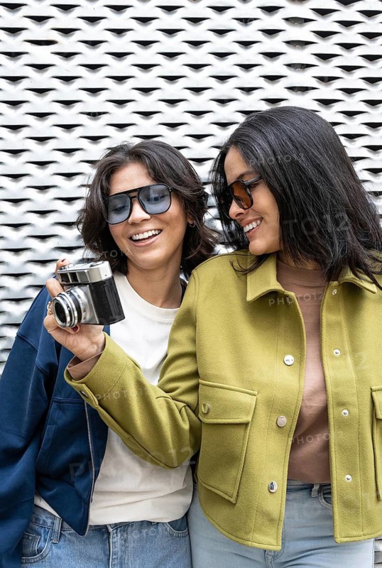 Two women share a joyful moment, posing with a vintage camera. They are smiling and wearing casual jackets, standing against a textured wall, conveying love and happiness