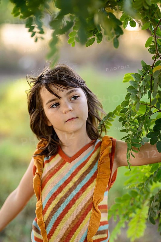 Young girl in a colorful striped outfit stands under a lush green tree. Her gaze is thoughtful, capturing a moment of quiet connection with nature