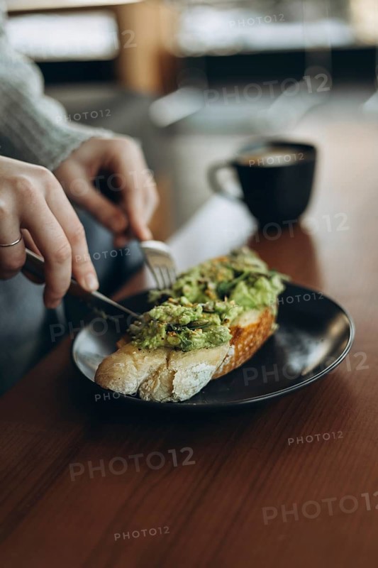 A cozy breakfast scene featuring a person preparing avocado toast on a dark plate, accompanied by a black cup of coffee, set on a wooden table in a relaxed atmosphere