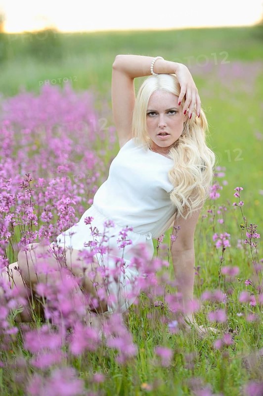 A woman with blonde hair poses dynamically among purple flowers in a grassy field, wearing a white dress