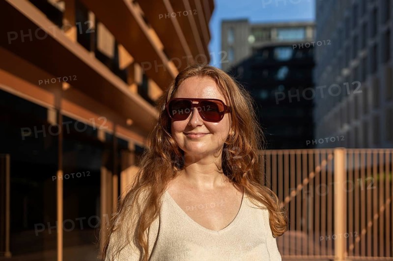 A woman with long hair and sunglasses enjoys the warm sunlight against an urban backdrop. Her relaxed expression and natural style reflect a modern, carefree lifestyle