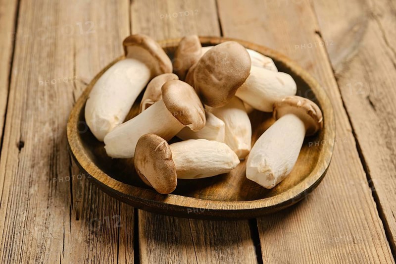 A round wooden plate filled with king oyster mushrooms. The scene shows a wooden table surface with a simple kitchen background. The mushrooms are clean and ready for cooking or serving in a dish