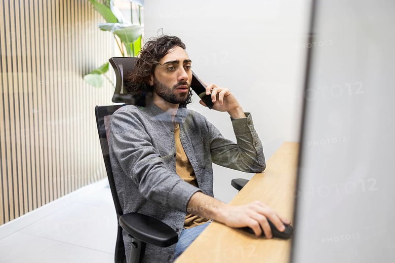 A focused professional sits at a sleek office desk, multitasking with a phone call while using a computer. The scene captures modern work dynamics in a bright office