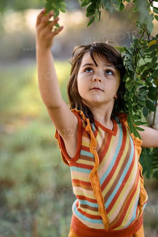 A young child reaches for vibrant green leaves in a sunny outdoor setting, wearing a colorful striped outfit. The scene captures innocence and wonder amidst nature