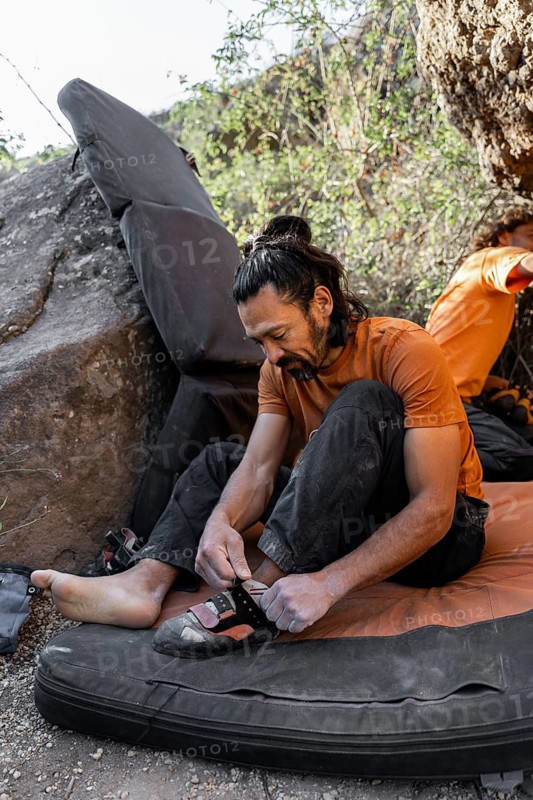 A climber prepares for a bouldering session by adjusting his climbing shoes on a crash pad, surrounded by natural rock formations and greenery, ready for an outdoor adventure