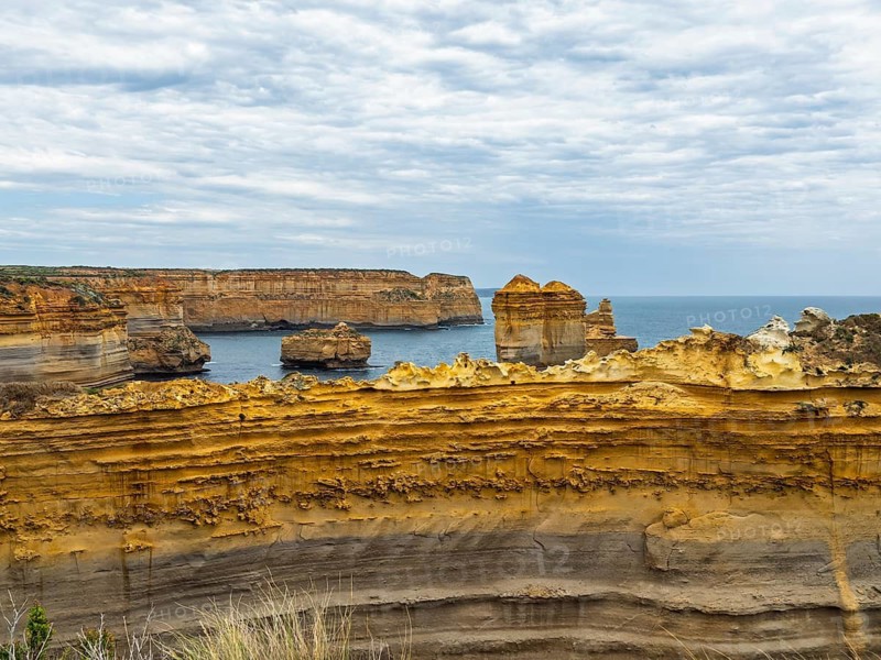 Stunning coastal rock formations under a cloudy sky in the Twelve Apostles, Australia. Layers of textured rock create a dramatic foreground, with the ocean and rugged cliffs in the distance