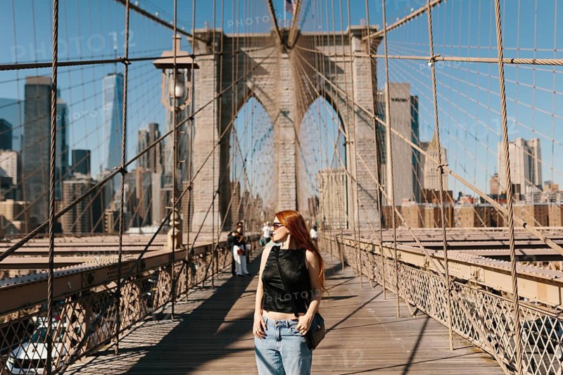 A woman with red hair walks across the Brooklyn Bridge, basking in the sun. The iconic bridge intricate cables and New York City skyline provide a stunning backdrop