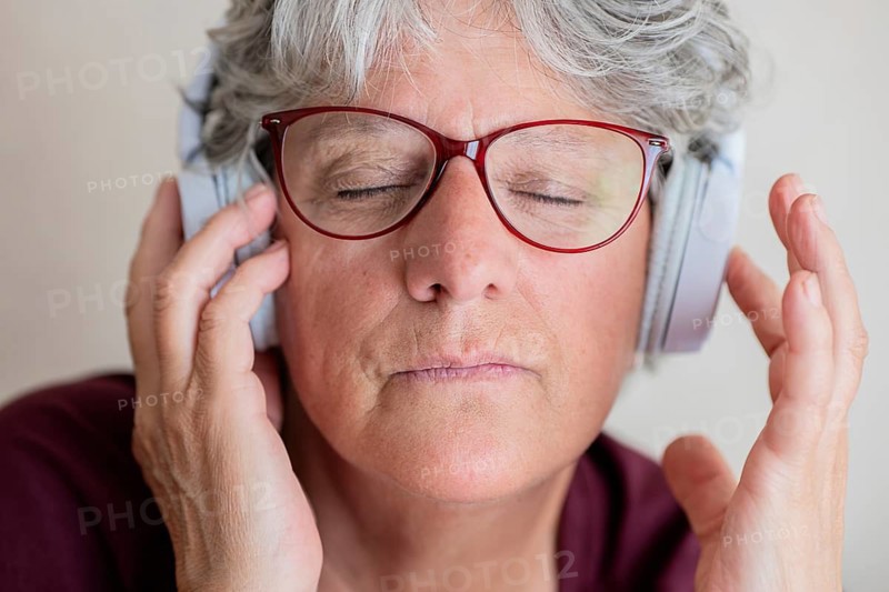 An senior woman with red glasses and silver hair enjoys music with closed eyes, fully immersed in the sound from her headphones. Her facial expression shows peace and relaxation