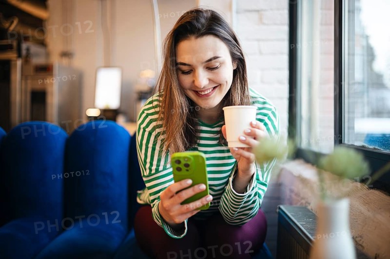 A smiling woman enjoys a cup of coffee as she engages with her smartphone in a cozy cafe setting