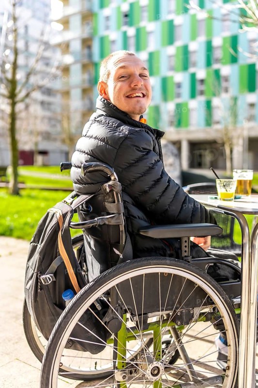 Happy disabled man sitting in a wheelchair, enjoying a refreshing drink at an outdoor table in an urban setting