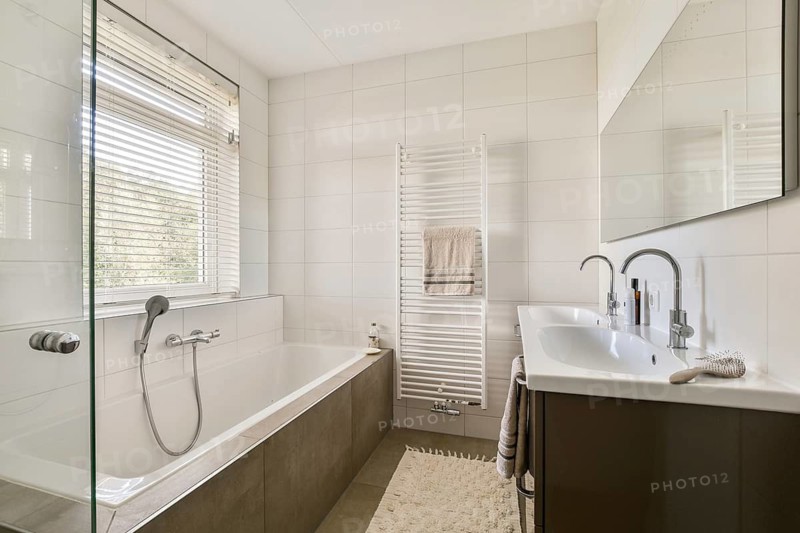 Bright and spacious modern bathroom featuring a large bathtub, sleek dual sinks, and a convenient towel heater. Natural light streams through the window with blinds, enhancing the serene ambiance