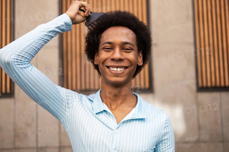 A cheerful non-binary individual with an afro hairstyle beams against an urban backdrop. The person, of mixed ethnicity, embraces their identity while showcasing confidence and joy