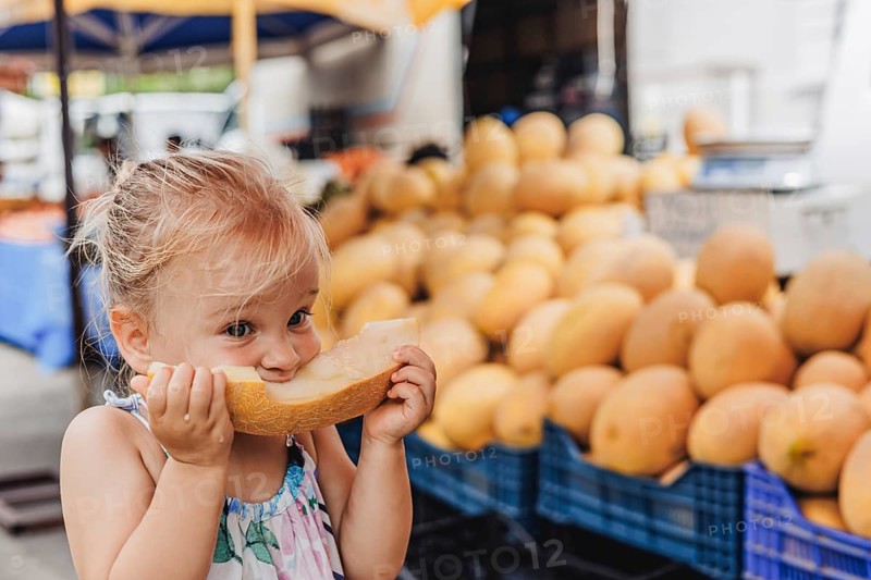 A young child contentedly bites into a slice of melon at a bustling local bazaar, surrounded by fresh produce, capturing the joy and simplicity of childhood moments