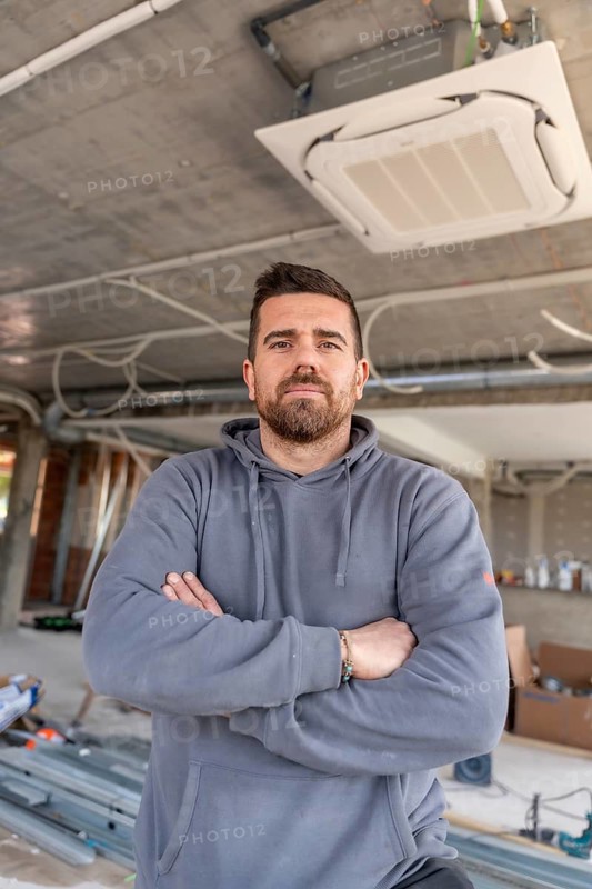 Hvac technician standing with arms crossed at a construction site, overseeing the installation of a new ceiling mounted air conditioning unit and other building systems