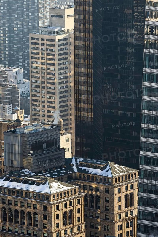 From above, this image captures a mix of modern and historic buildings in New York, showcasing contrasting architectural styles. The view highlights a variety of building facades and textures under a clear sky