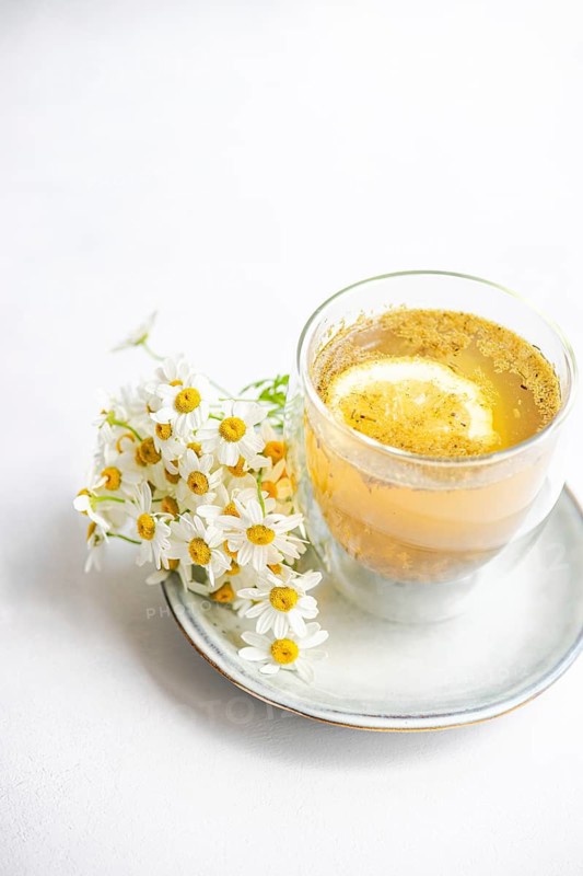 A serene still life featuring bright summer daisies next to a glass of lemon tea on a white ceramic plate. The simple and elegant setup is against a soft white background