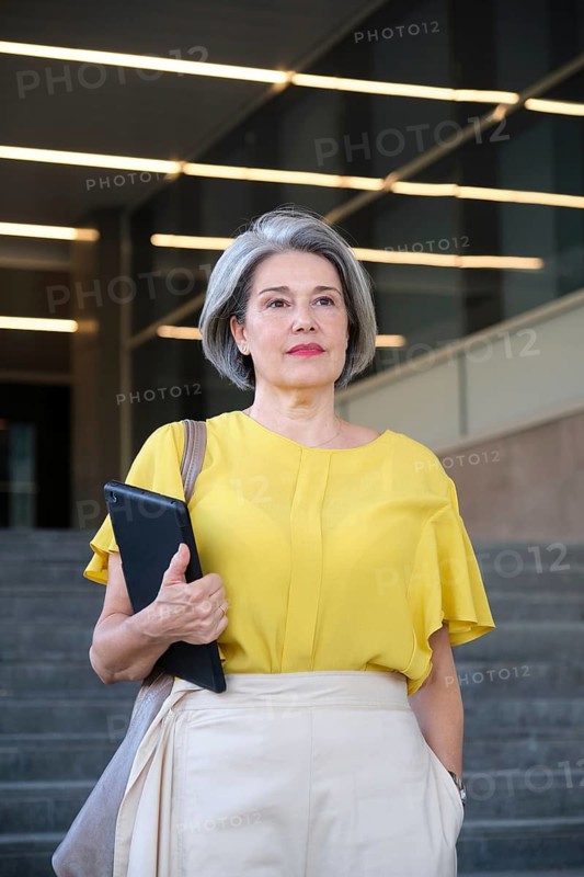 Mature woman standing confidently outdoors, wearing a stylish yellow top, holding a tablet. She exudes professionalism and modernity in an urban environment