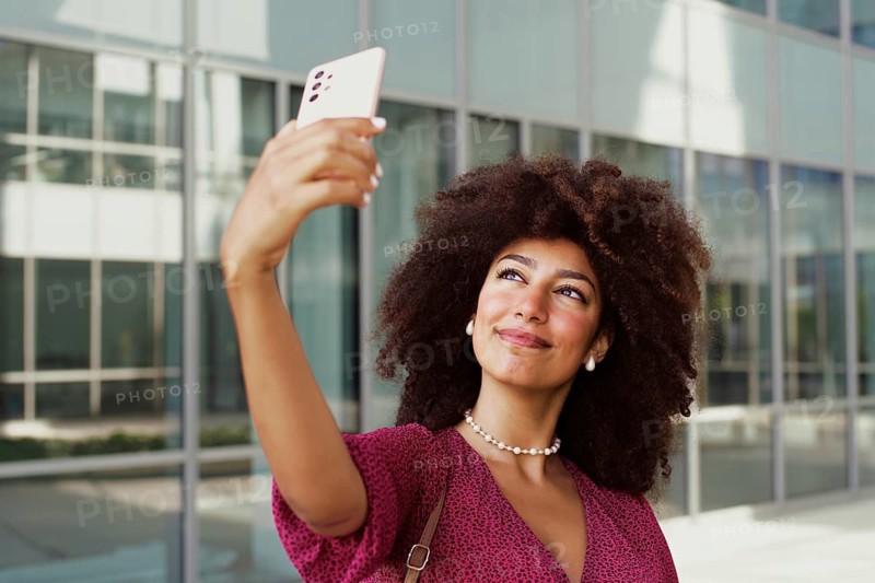 A young Arab woman with curly hair takes a selfie with her smartphone outside a modern glass building, capturing a joyful moment in the bright sun