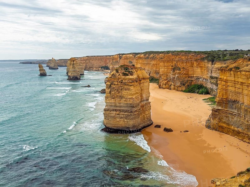 Stunning coastal cliffs and rock stacks rise dramatically from the ocean, set against a backdrop of rolling waves and sandy shores under a cloudy sky in the Twelve Apostles, Australia