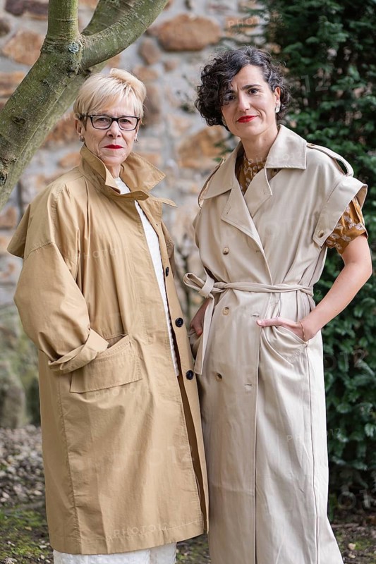 Two fashion women in stylish trench coats pose outdoors. They exude confidence and elegance amid a natural setting, creating a blend of fashion and nature