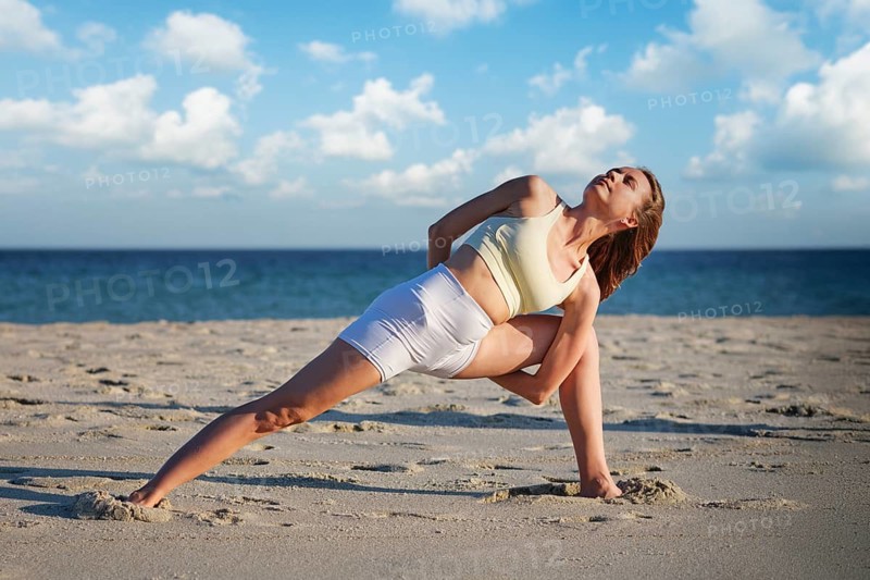 A woman practices Baddha Trikonasana with legs wide apart on sandy beach during sunset, arms bound behind her back in yoga pose
