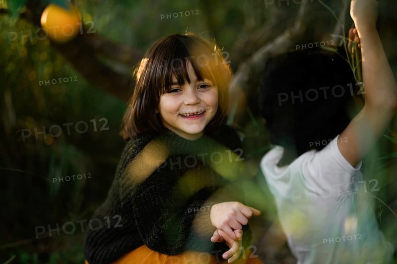 Joyful children with dark hair smiles warmly while playing outdoors on a sunny day, surrounded by lush greenery and orange trees, conveying happiness and innocence
