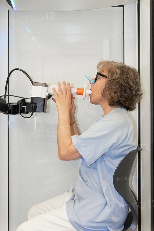 A female patient is shown performing a spirometry test in a clinical environment to assess her pulmonary function. She wears a nasal clip and uses a mouthpiece connected to the spirometer