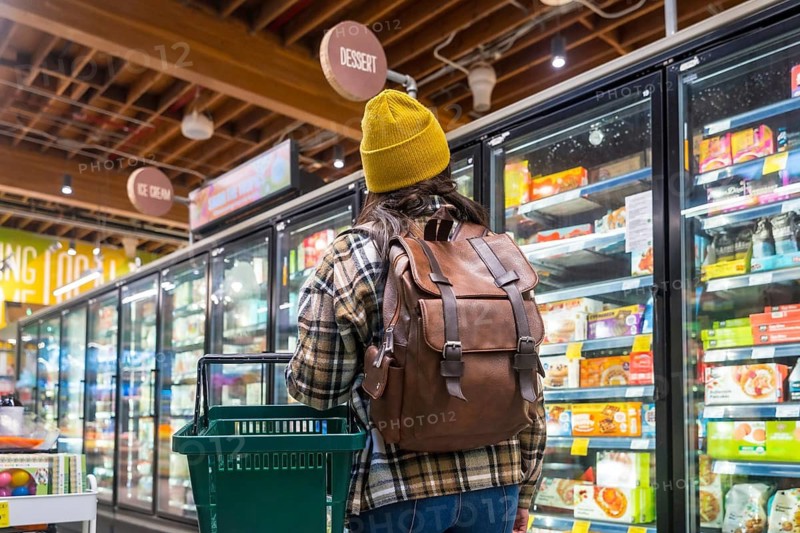 Woman in beanie and plaid shirt with backpack and shopping basket browsing frozen food in a modern supermarket freezer aisle, choosing items for meals and snacks during everyday shopping