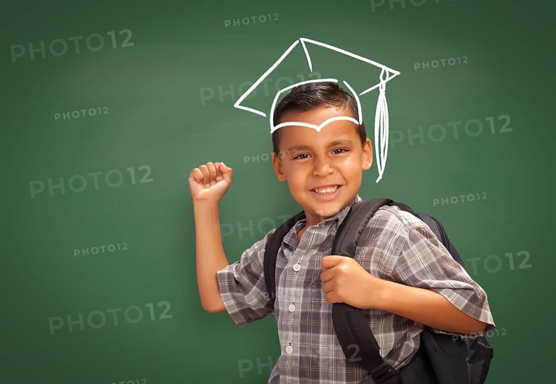 Young Hispanic Student Boy Wearing Backpack Front Of Blackboard with Graduation Cap Drawn In Chalk Over Head