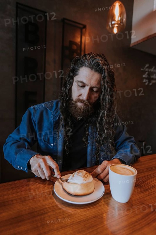 A bearded man sits in a cozy cafe enjoying a hot coffee and a pastry. The ambient lighting and casual attire create a relaxed and inviting atmosphere