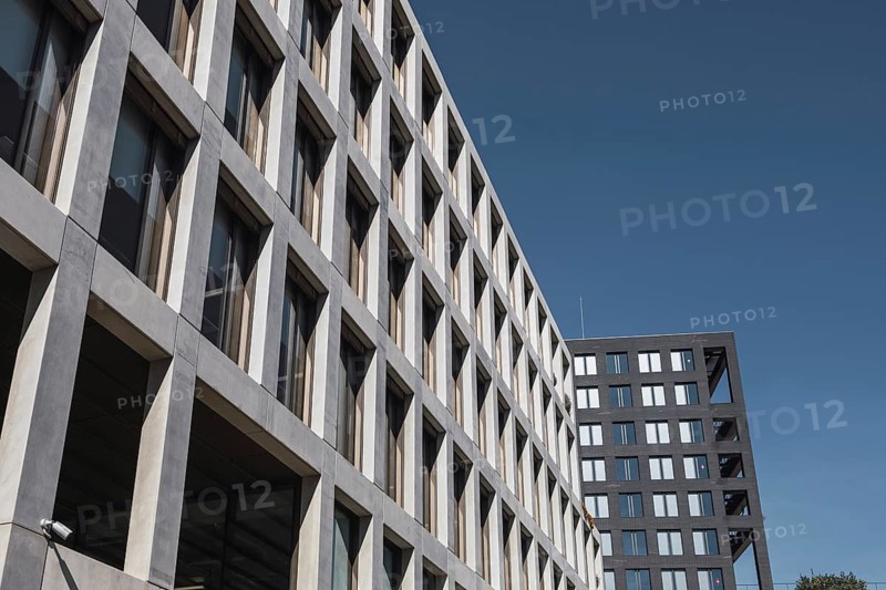 A perspective view of modern urban office buildings with geometric designs against a clear blue sky. Architectural details highlight symmetry and contemporary aesthetics in an urban setting