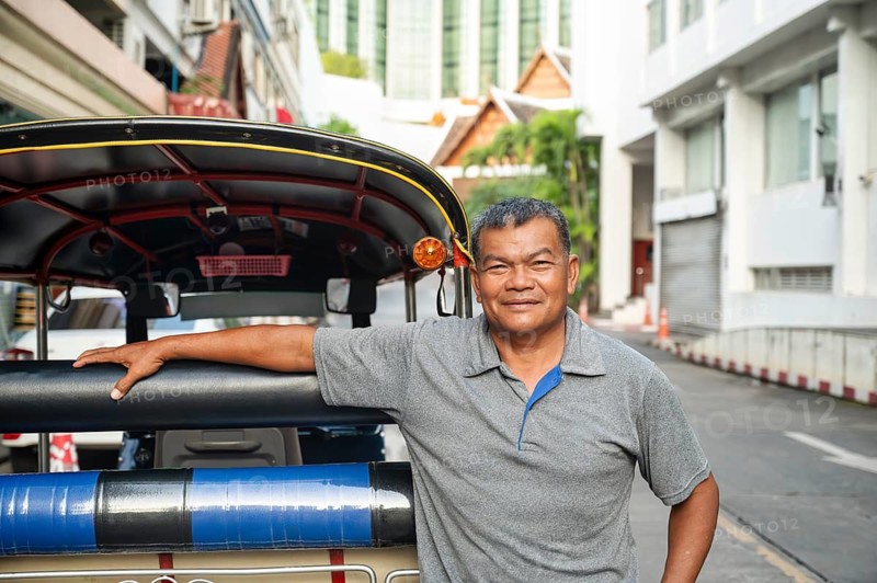 A Thai tuk tuk driver stands confidently with his arm resting on his colorful vehicle, smiling in the streets of Chiang Rai, Thailand