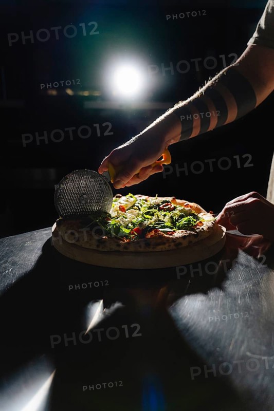 A hand with a tattoo slices a gourmet pizza topped with arugula, cheese, and cherry tomatoes using a pizza cutter, dramatically lit against a dark background