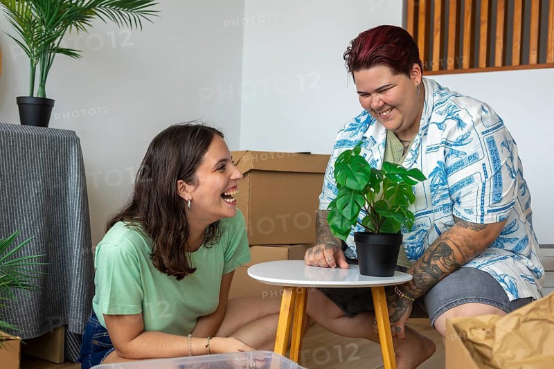Lesbian friends are joyfully arranging a potted plant on a small table in a cozy indoor setting. Surrounded by boxes, they share laughter and create a warm, inviting atmosphere