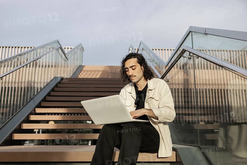 A man sits on outdoor steps, focused on his laptop He embraces a modern, flexible working lifestyle, utilizing technology in an open, urban environment under clear skies