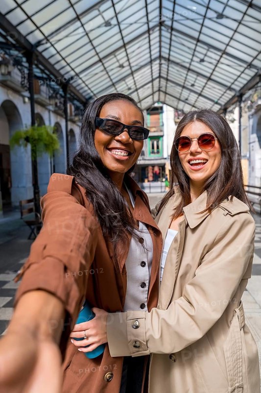 Two cheerful businesswomen hugging and taking a selfie while walking in a shopping mall with glass roof
