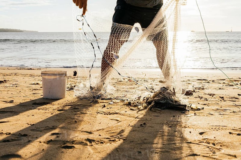 A fisherman stands on a sunlit beach, collecting his catch from a fishing net. Shadows stretch across the sand, and the ocean waves gently lap the shore in the background