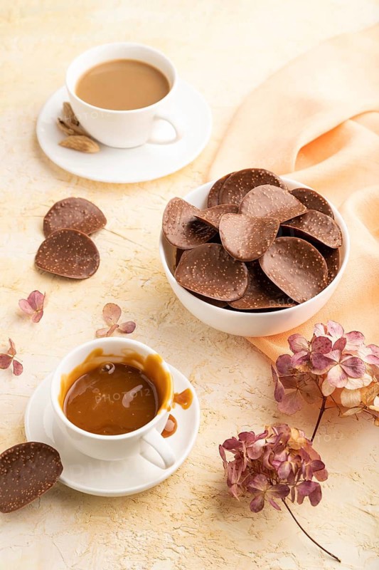 Chocolate chips with cup of coffee and caramel on a beige concrete background and orange textile. side view, close up