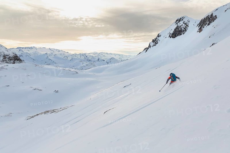 A lone ski tourer skis down a pristine, snow-covered mountain slope under a bright, cloud-streaked sky, showcasing the serene and adventurous nature of ski touring