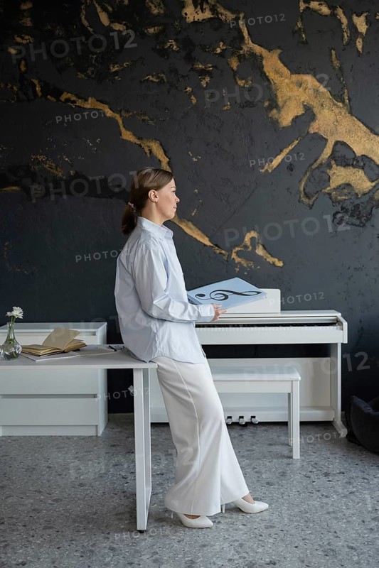 Woman sitting at a table with a piano and open music book, preparing piano lessons. The setting is a modern, stylish room with a focus on musical education