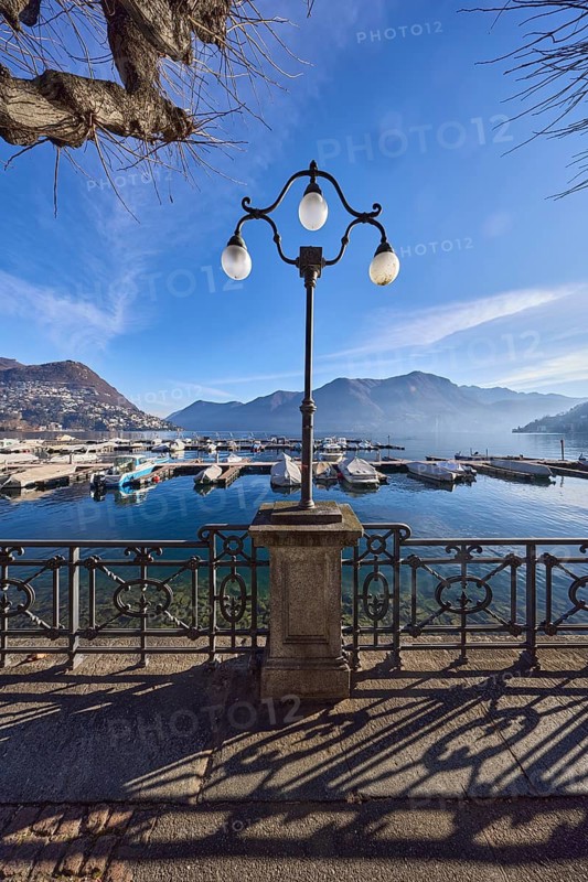 Lantern, forged railing, lakeside promenade, shadow, marina, trees, motorboats, hill with village, haze, backlight, blue sky with veil clouds, Lake Lugano, Lugano, district Lugano, canton Ticino, Switzerland