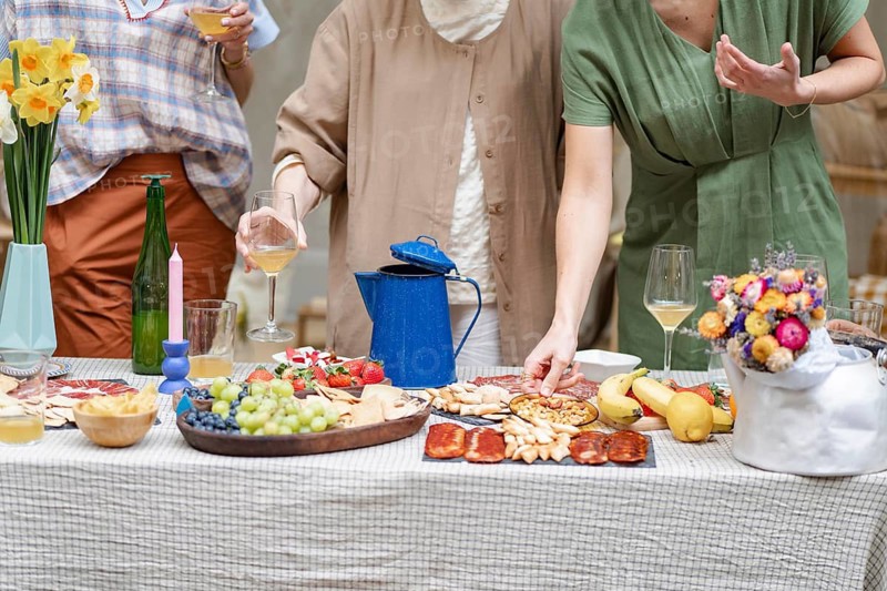 A casual gathering features coworkers enjoying wine and appetizers around a decorated table. Vibrant flowers and a variety of snacks create a warm, inviting atmosphere