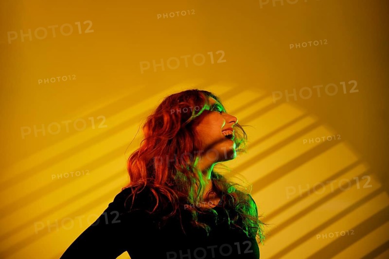 A young cheerful woman with flowing hair is surrounded by vibrant green and yellow studio lights. Shadows create dynamic patterns against the bright backdrop, evoking a sense of motion and energy