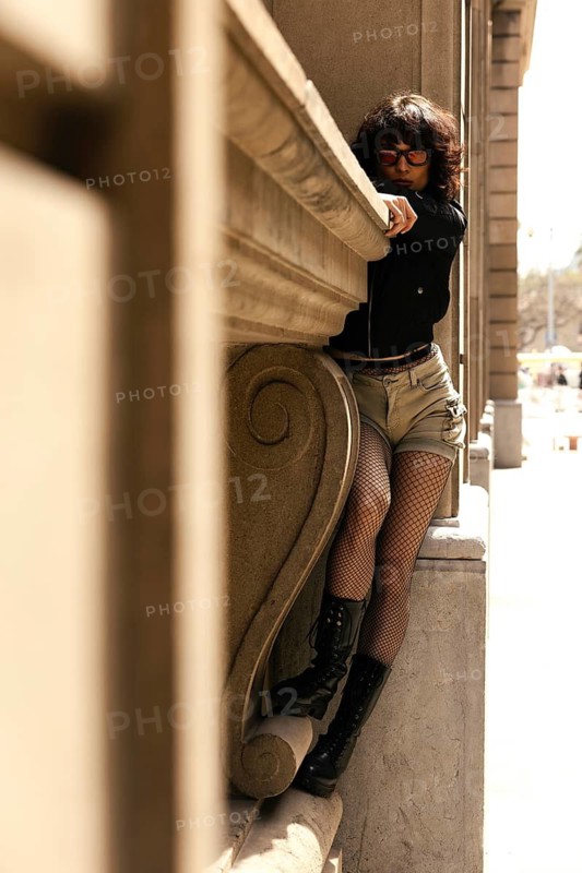 A stylish woman with curly hair and sunglasses leans against a classic stone structure. Dressed in fishnet tights, boots, and a cropped jacket, she exudes confidence, attitude, and urban edge