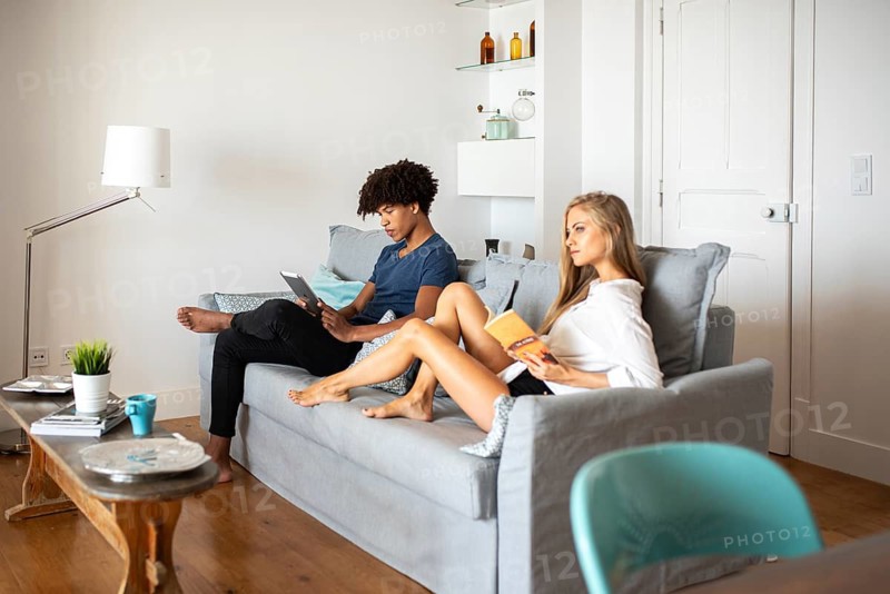 Couple relaxing in a bright, modern living room. One reads a book while the other uses a tablet. They sit on a gray sofa, surrounded by minimalistic decor
