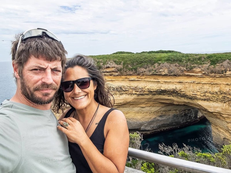 Two people smiles at the camera, enjoying a bright day by the ocean in the Twelve Apostles, Australia. They stand in front of a rugged cliff formation with lush green foliage
