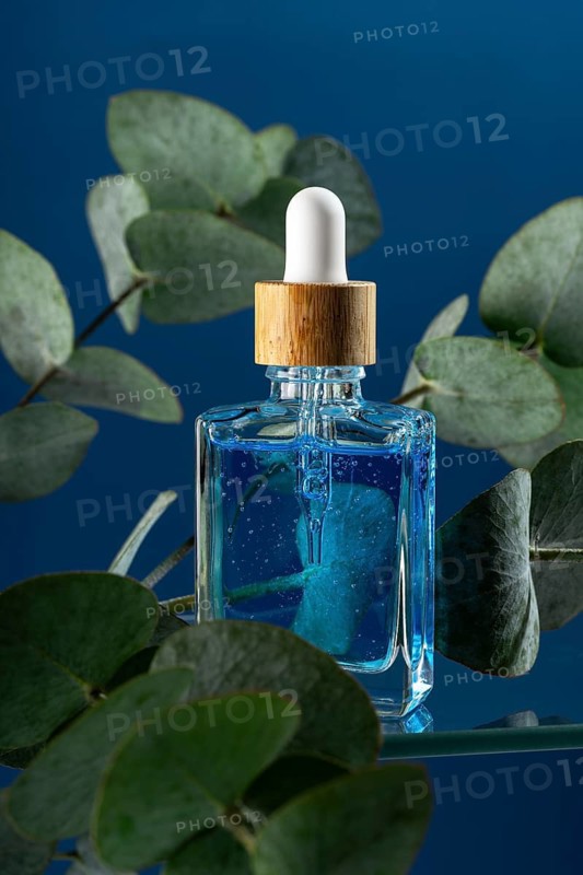 A cooling blue face care serum or oil targeted for men, displayed in an elegant dropper bottle surrounded by eucalyptus leaves on a dark blue background. Ideal for skincare routines