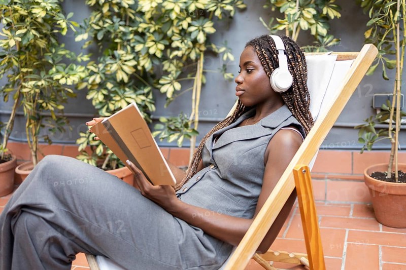 A young African woman with braids listens to music on headphones while relaxing with a notebook in a sunny coworking space, surrounded by lush plants and natural light